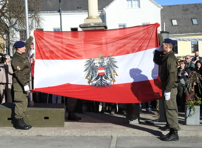 A man in a military uniform holds a large Austrian flag in front of a monument as a crowd watches.