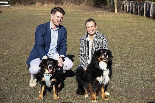 Ein Mann und eine Frau sitzen auf einem Grasfeld mit zwei Hunden und lächeln für die Kamera.