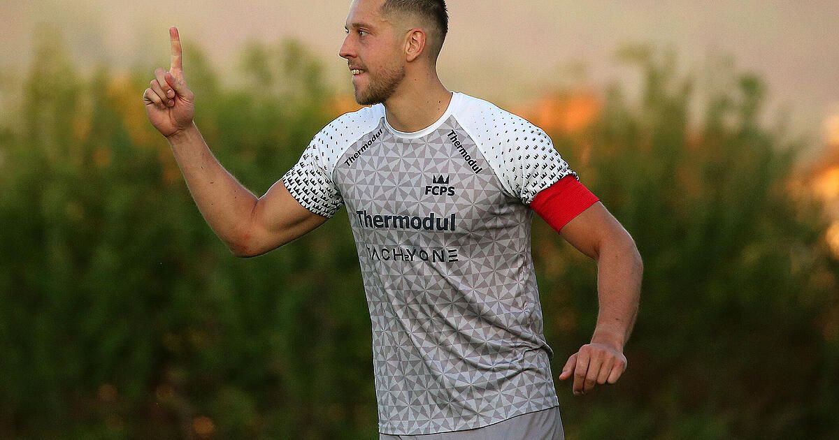 Ein Fußballspieler zeigt mit einem Finger nach oben, möglicherweise um ein Tor zu feiern. Er trägt ein graues und weißes Thermomodul-Trikot mit roten Ärmeln.
