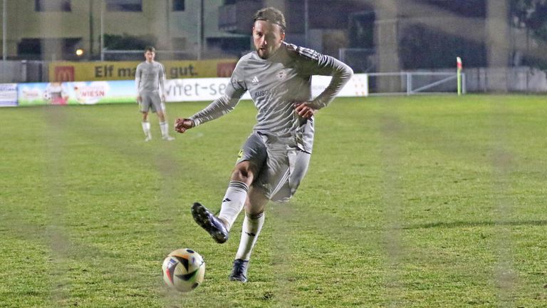 A soccer player in a gray uniform kicks a ball on a grassy field. In the background, another player is standing.