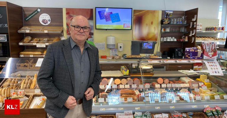 Ein Mann steht in einem Geschäft, hinter einer Vitrine mit Fleisch und Käse. Ein Monitor zeigt einen Preis und ein gelbes Käseposter an, der an der Wand montiert ist.