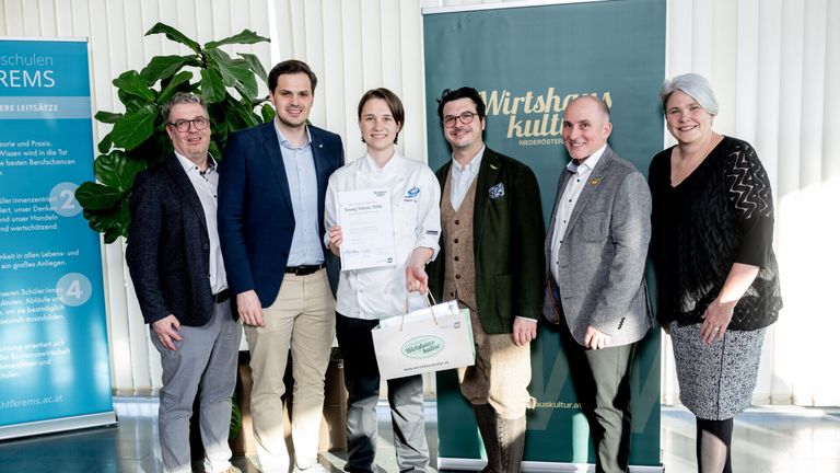 Fünf Personen stehen vor einem Banner, halten einen Papierbeutel und ein Zertifikat. Die zentrale Figur, eine Frau in einer Kochuniform, lächelt und hält ein Zertifikat. Der Mann auf ihrer linken Seite trägt einen dunklen Anzug. Der Mann auf ihrer rechten Seite trägt einen grünen Mantel.