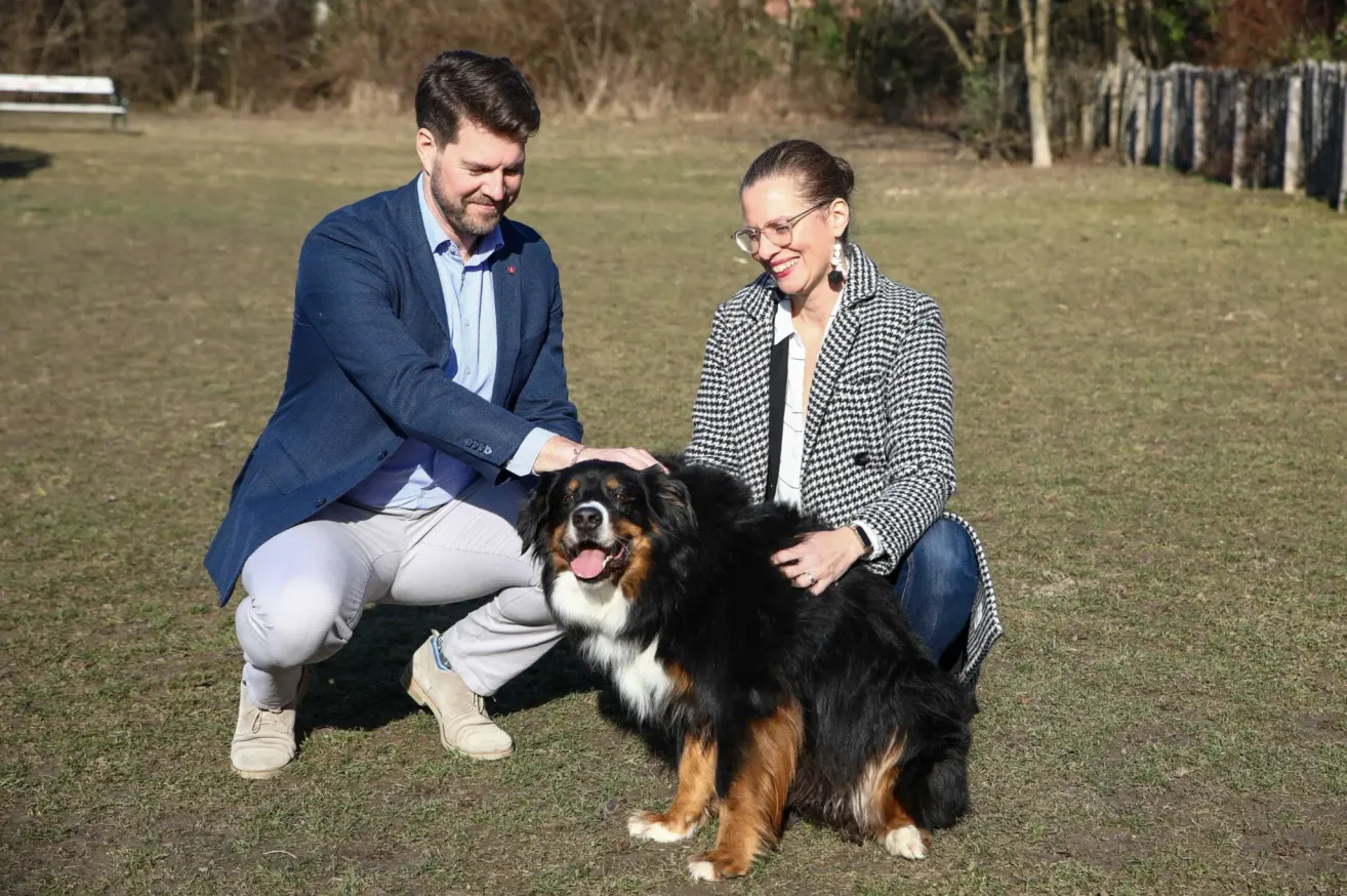 Ein Mann und eine Frau knien lächelnd auf dem Gras und streicheln einen Berner Sennenhund. Der Mann trägt einen blauen Blazer und weiße Hosen, während die Frau einen schwarz-weiß karierten Mantel trägt. Der Hund sitzt fröhlich zwischen ihnen.