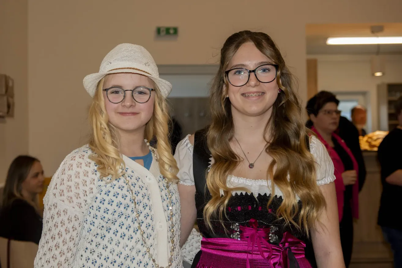 Two girls stand together; one in a white dress with a hat, the other in a pink and black outfit. Both are smiling for the camera.