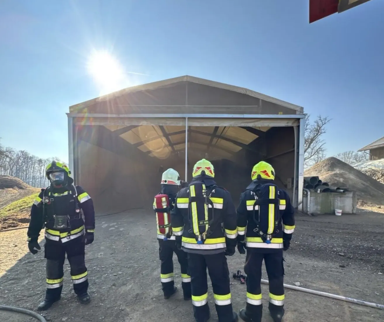 Vier Feuerwehrleute stehen vor einem Gebäude. Sie tragen Schutzanzüge und Helme. Ein Feuerlöscher ist an einem ihrer Rücken montiert. Das Gebäude hat ein Metalldach und Glasfenster.