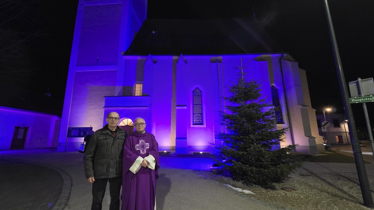 Zwei Männer stehen vor einer Kirche mit lila Beleuchtung. Ein Mann trägt eine Brille und eine schwarze Jacke. Der andere Mann ist in einen violetten Umhang mit einem Kreuz auf der Vorderseite gekleidet.