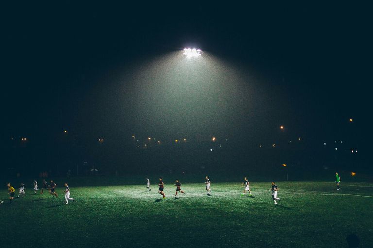Ein Nachtfußballspiel mit Spielern, die auf einem Feld unter einer Flutlichtstrahler laufen, mit einem dunklen Himmel und entfernten Stadtlichtern.