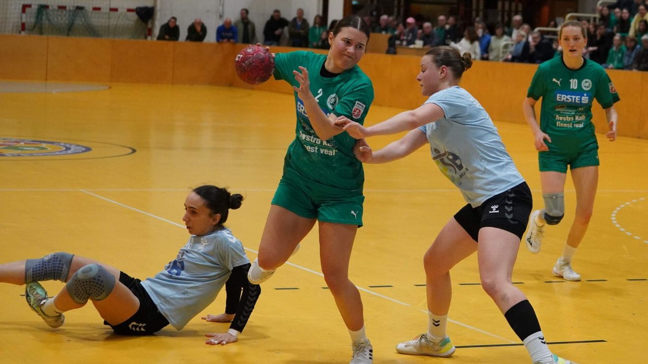 Drei weibliche Spielerinnen auf einem Spielfeld, die ein Ballspiel spielen, eine hält einen Ball, eine andere verteidigt und die dritte liegt auf dem Boden.