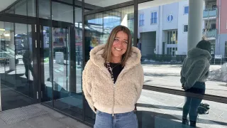 Eine lächelnde Frau in einer weißen Jacke und Jeans steht vor einem Gebäude mit Glaswänden. Hinter ihr ist die Reflexion einer weiteren Person in einer Pelzjacke zu sehen.