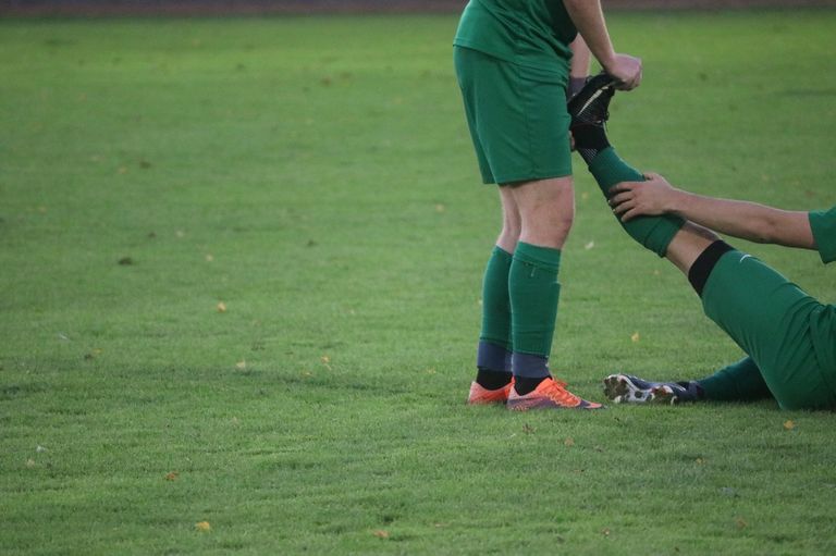 A soccer player in green uniform assists another player who is injured on the field, both wearing cleats.