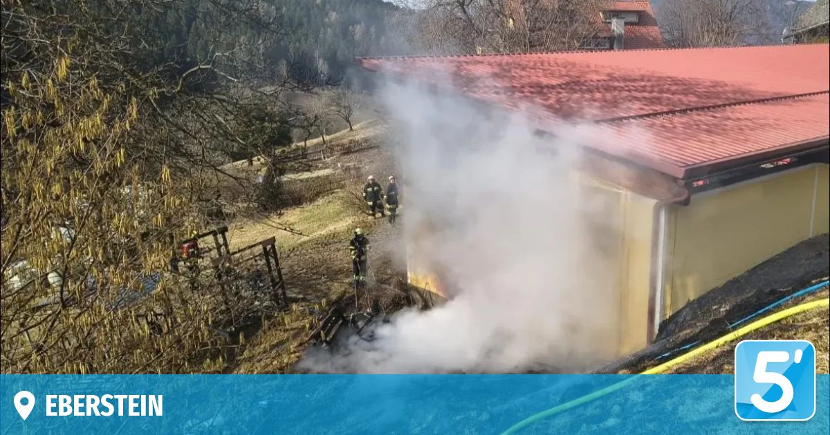 Ein Haus mit rotem Dach steht in Flammen, Rauch steigt auf. Feuerwehrleute sind anwesend und stehen um das Haus herum.