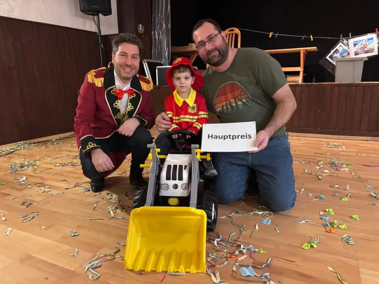 Drei Männer knien auf einem Holzboden mit einem Spielzeugbagger. Der Mann in der Mitte hält ein Schild, das Hauptpreis sagt. Der Mann auf der linken Seite trägt ein Kostüm.