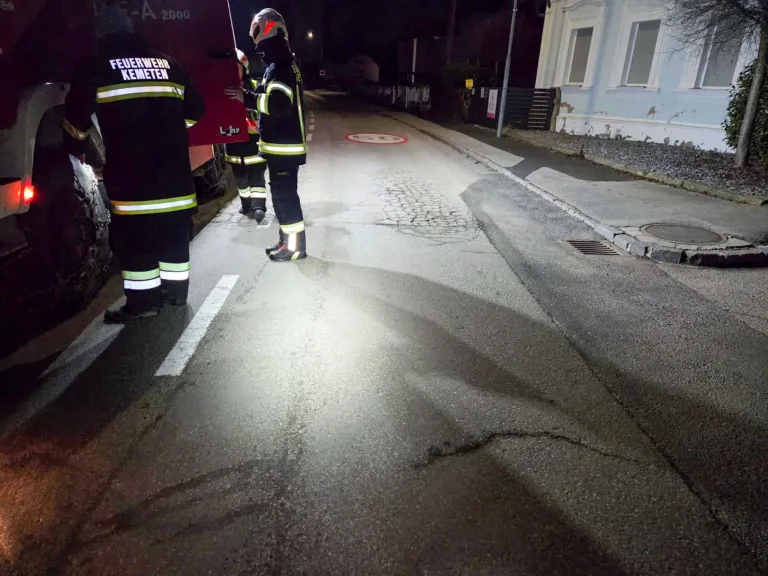 Firefighters in protective gear stand on a road, illuminated by their vehicle's headlights. A curb and building are visible.