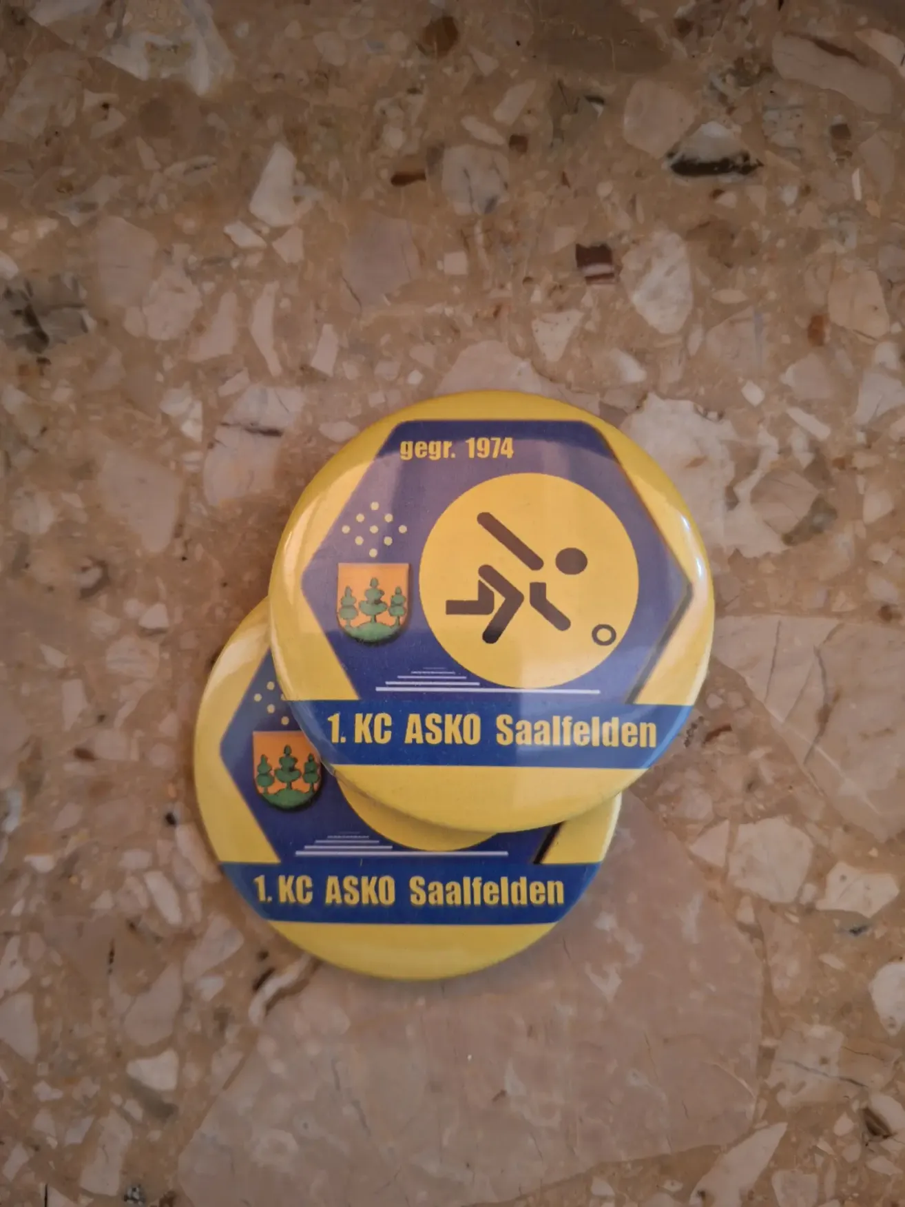 Zwei gelbe runde Anstecker mit blauen Rändern sind aufeinandergestapelt. Sie tragen den Text '1. KC ASKO Saalfelden' und ein Logo mit einer gehenden Figur und dem Jahr '1974.'