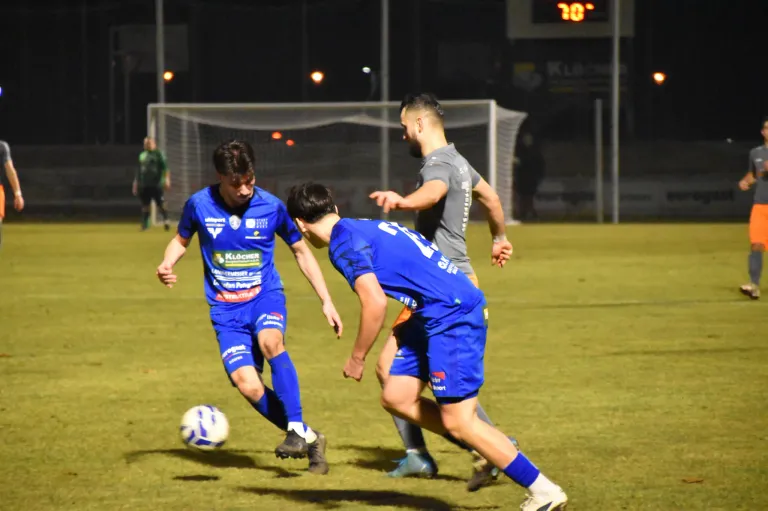 Drei Fußballspieler spielen bei Nacht auf einem Feld. Zwei tragen blaue Trikots, der andere grau. Sie jagen dem Ball hinterher, der auf dem Boden liegt.