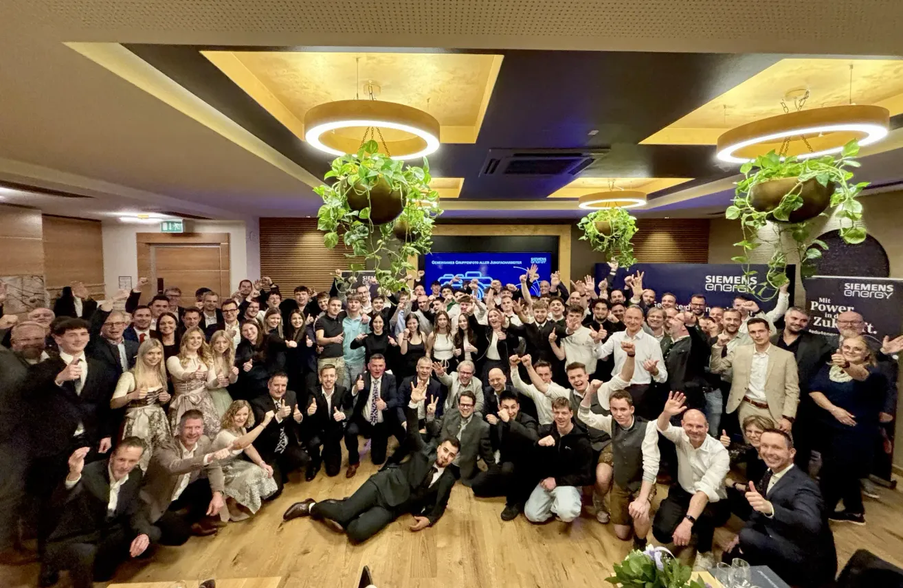 Eine Gruppe von Menschen versammelt sich für ein Foto, einige stehend und andere auf dem Boden sitzend. Der Raum hat eine moderne Einrichtung mit hängenden Pflanzen und einer großen Leinwand an der Wand.