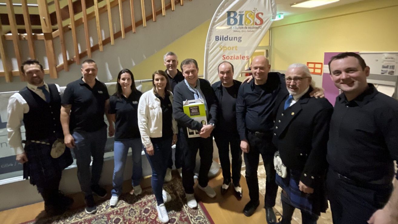 Eine Gruppe von Menschen steht drinnen und posiert für ein Foto. Ein Banner hinter ihnen sagt BISS. Sie sind feierlich gekleidet. Der Mann in der Mitte hält eine Box.