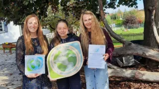Drei Frauen stehen lächelnd unter einem Baum, halten ein Schild und ein Zertifikat. Die mittlere Frau hält ein Schild mit einer Erde und den Worten 'Wir sind Klimabündnis'. Sie scheinen eine Errungenschaft zu feiern.