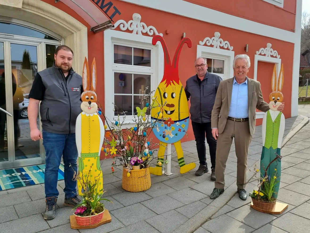 Vier Männer stehen vor einem Gebäude mit großen Hasenfiguren. Die Männer lächeln und einer berührt einen Hasen. Die Hasen sind in leuchtenden Farben mit Blumen bemalt.