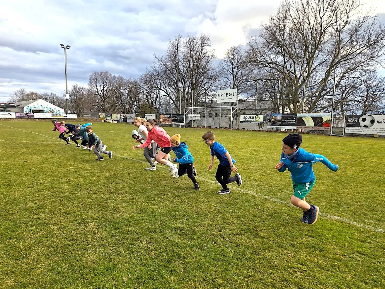 Eine Gruppe von Kindern in Turnschuhen läuft auf einem Rasenfeld unter einem bewölkten Himmel. Im Hintergrund steht ein Gebäude und Bäume.