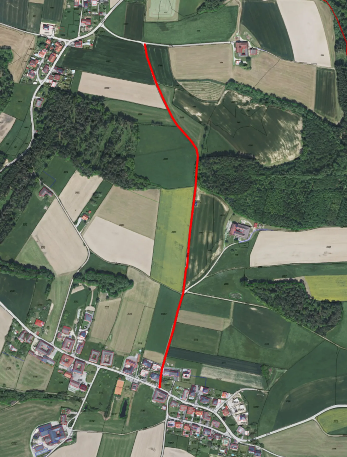 Eine Luftaufnahme eines ländlichen Gebiets mit mehreren Gebäuden, Straßen und Feldern. Eine rote Linie markiert einen bestimmten Weg oder eine Route durch die Landschaft.