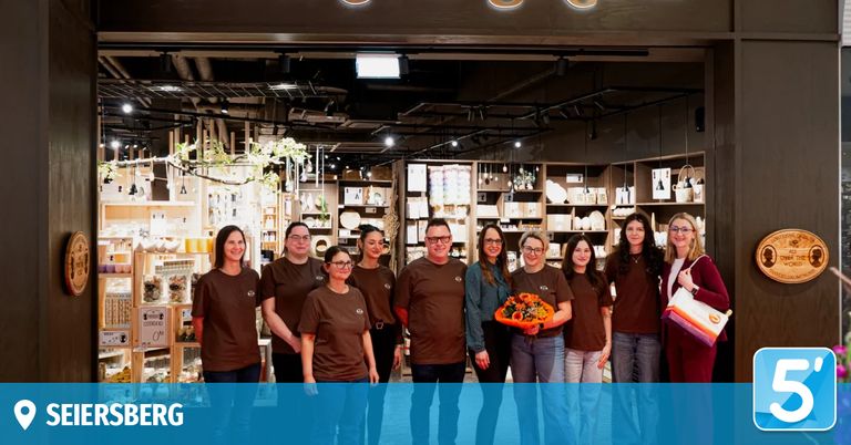 Eine Gruppe von Menschen in braunen T-Shirts steht in einem Geschäft, lächelt und posiert für ein Foto. Eine Frau hält einen Blumenstrauß. Regale mit Produkten sind hinter ihnen.