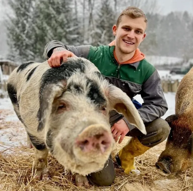 Ein Mann in einer grünen Jacke hockt im Schnee mit zwei Schweinen, von denen eines vor ihm steht.