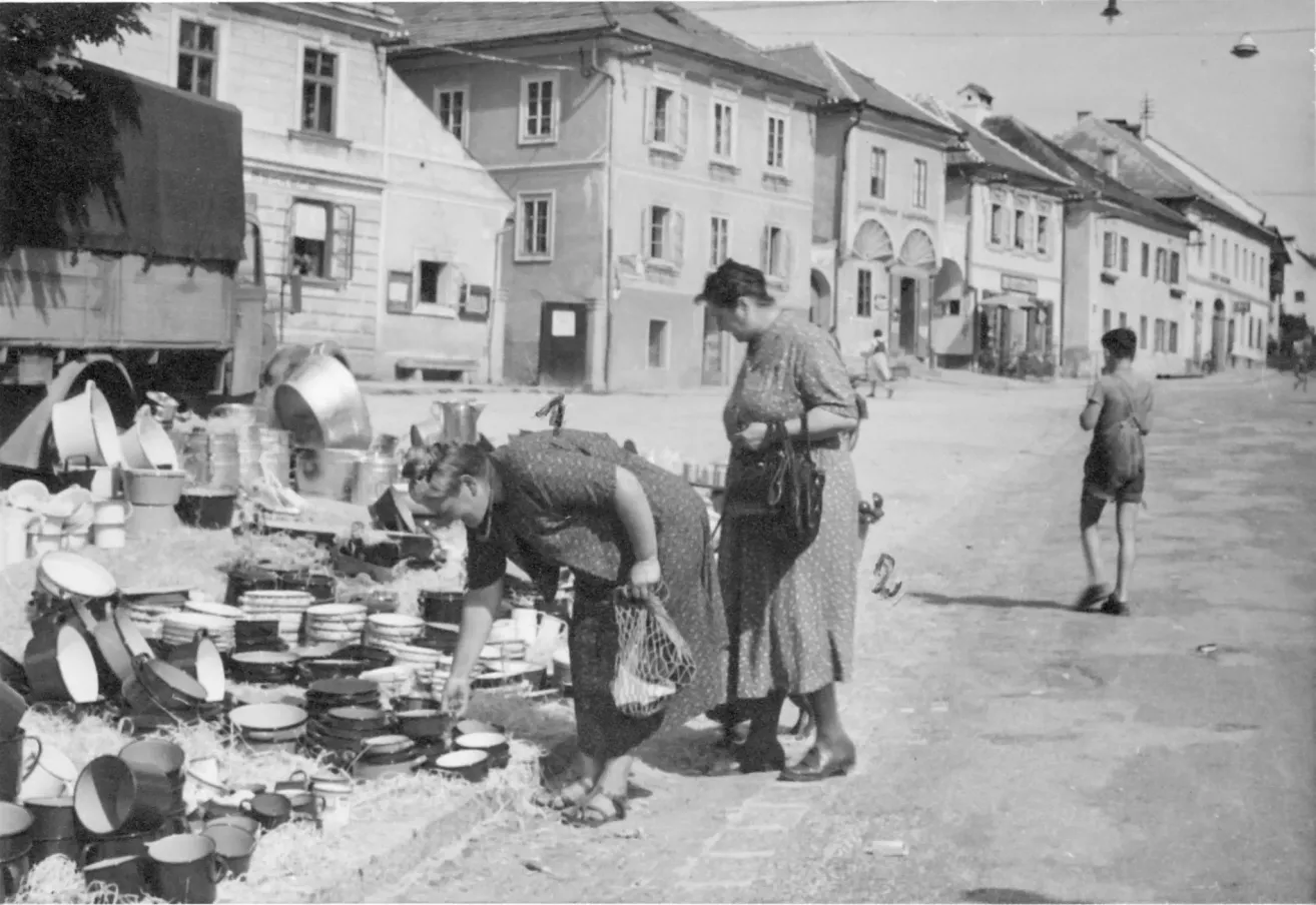 Ein Schwarz-Weiß-Foto von zwei Frauen, die in einem Markt mit Stapeln von Kochgeschirr stehen. Gebäude mit vielen Fenstern säumen die Straße.