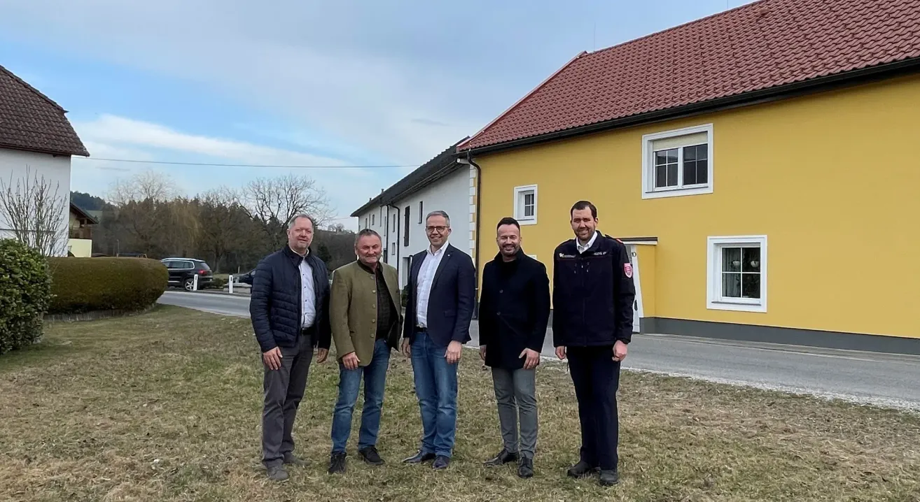 Fünf Männer stehen vor einem gelben Haus mit einem roten Dach. Sie tragen formelle Kleidung, möglicherweise bei einer Veranstaltung. Hinter ihnen hat das Haus weiße Fenster und einen schwarzen Schornstein.