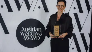 A woman in black is holding an award, smiling at the camera. She stands in front of a white backdrop with the text 'Austrian Wedding Award'.