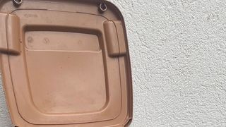 An open brown plastic bin with a label and a handle is positioned against a white textured wall.