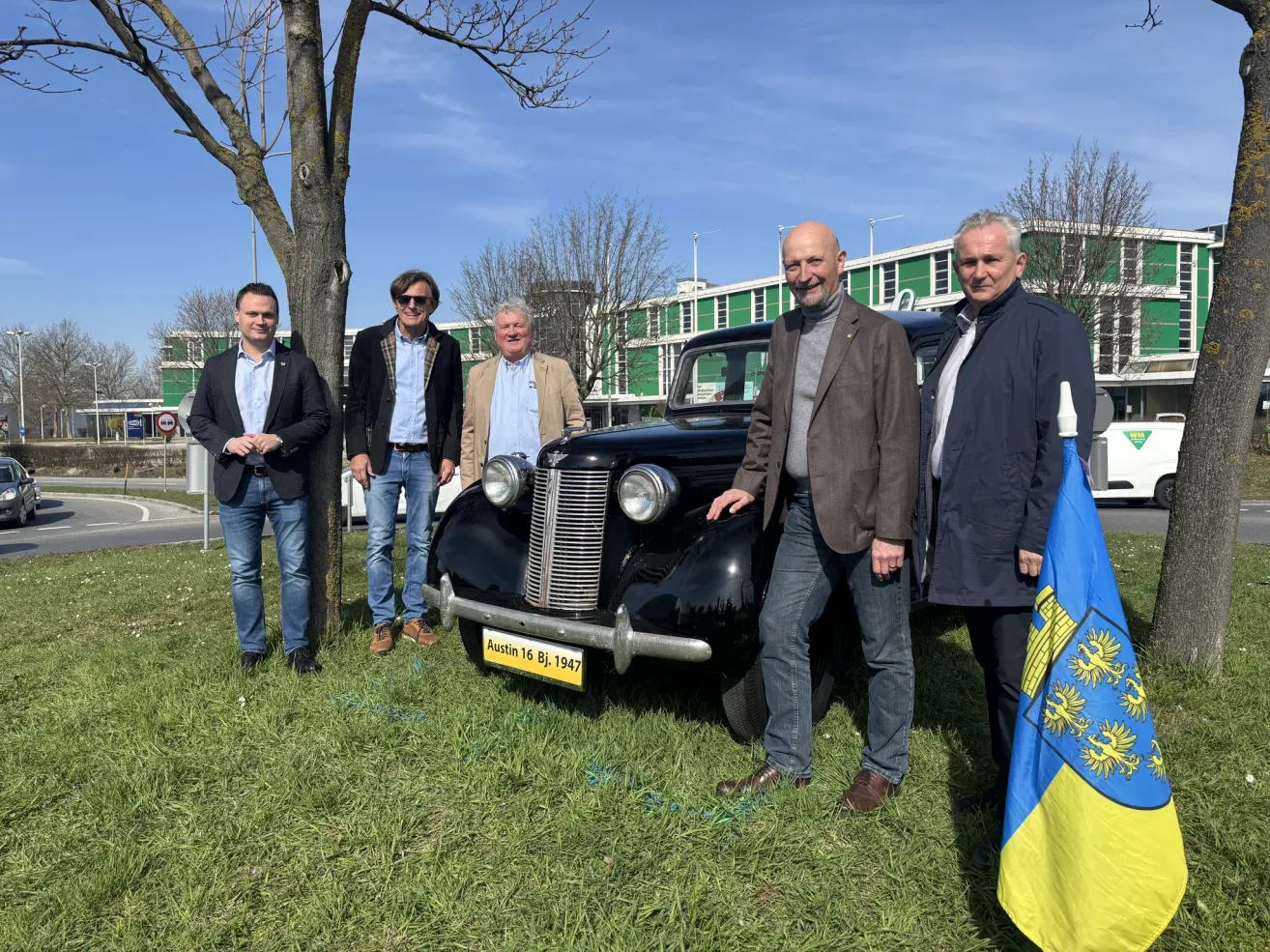 Fünf Männer stehen neben einem klassischen schwarzen Auto mit dem Kennzeichen Austin 16 BJ 1947. Sie alle lächeln und scheinen für ein Foto zu posieren. Sie stehen auf einem Rasen.