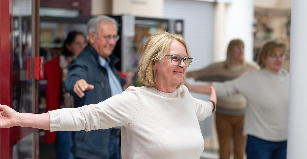 Eine ältere Frau mit Brille lächelt und streckt ihre Arme, während sie an einer Gruppenaktivität teilnimmt. Ein Mann hinter ihr schaut zu, und andere sind im Hintergrund verschwommen.