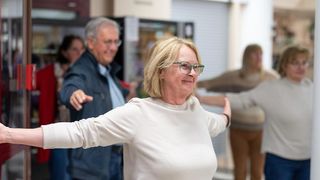 Eine ältere Frau mit Brille lächelt und streckt ihre Arme, während sie an einer Gruppenaktivität teilnimmt. Ein Mann hinter ihr schaut zu, und andere sind im Hintergrund verschwommen.
