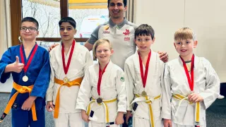 Eine Gruppe junger Jungen in Kampfsportuniformen mit Medaillen um den Hals posiert für ein Foto mit einem lächelnden Mann in einem grauen Shirt.