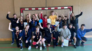 A group of students with medals are posing for a photo in front of a goal net. They are holding paddles and some are raising their hands.