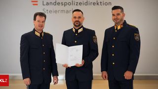 Drei Männer in Polizeiuniformen stehen vor einem Schild, auf dem 'Landespolizeidirektion Steiermark' steht. Der Mann in der Mitte hält ein Dokument.
