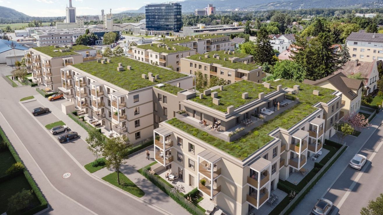 Luftaufnahme von modernen Gebäuden mit grünen Dächern, Balkonen und Außenbereichen. Bäume und Pflanzen umgeben den Komplex. Im Hintergrund befinden sich Gebäude, darunter ein großer Turm.