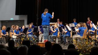 Ein Orchester spielt auf der Bühne. Die Bandmitglieder tragen blaue Hemden und sitzen auf Stühlen, während sie ihre Instrumente spielen. Eine Frau vor ihnen dirigiert.