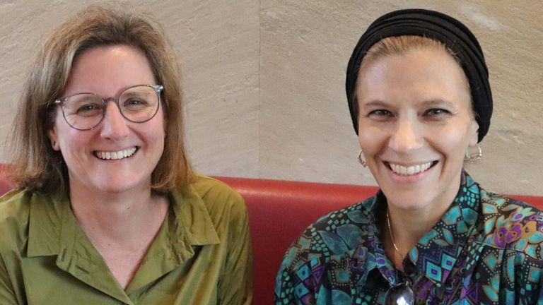 Zwei Frauen sitzen auf einer roten Bank und lächeln, eine trägt eine Brille und die andere eine Kopfbinde und eine Halskette.