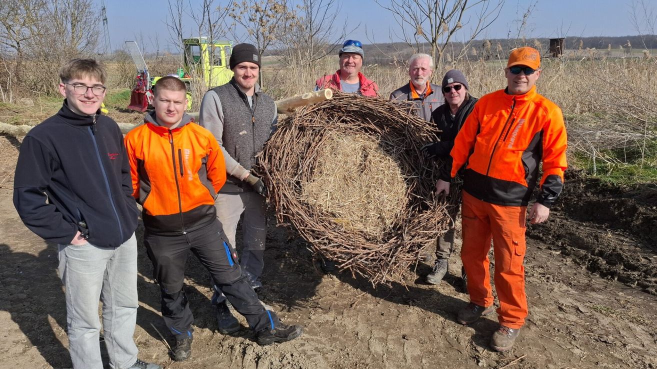 Fünf Männer in orangefarbenen Jacken und Mützen posieren mit einem großen Nest aus Zweigen und Heu auf einem Feld mit Bäumen und einem gelben Maschinenfahrzeug im Hintergrund.