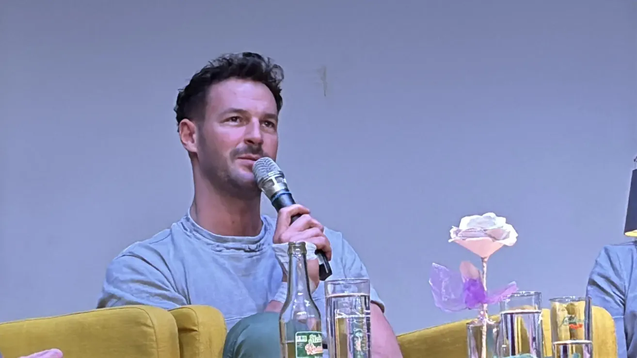 A man speaks into a microphone while seated in front of a yellow couch. A glass of water and a bottle are on the table next to him.
