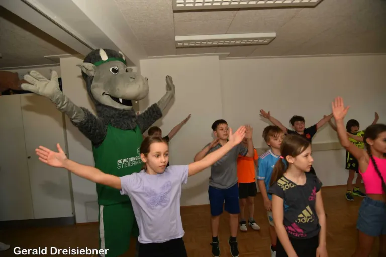 Ein Maskottchen in einem grünen Kostüm steht mit erhobenen Armen in einem Klassenzimmer mit Kindern. Sie alle machen eine Übung.