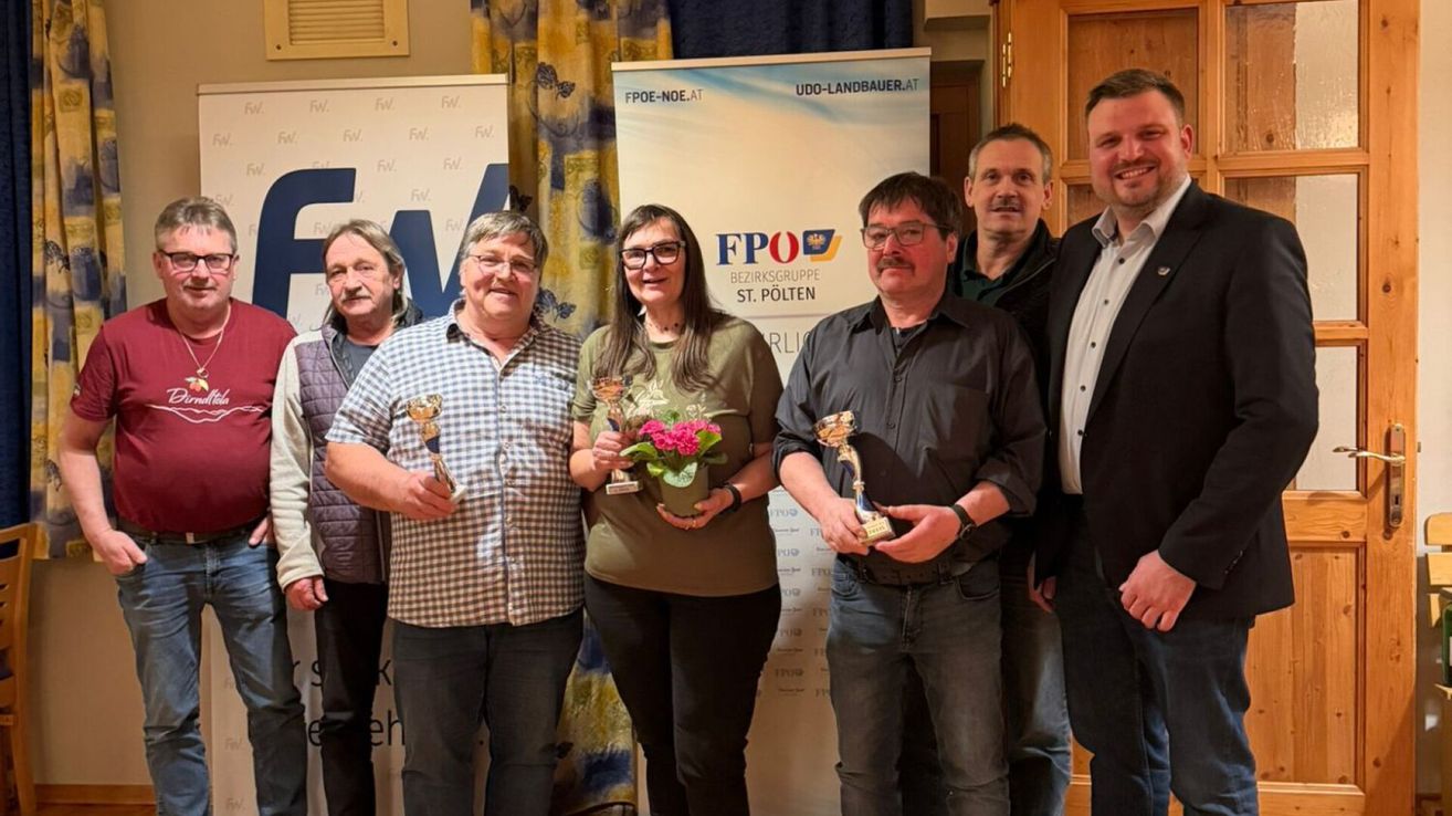 Sechs Menschen stehen zusammen und halten Trophäen. Ein Banner mit dem Text 'FPO' ist hinter ihnen sichtbar.