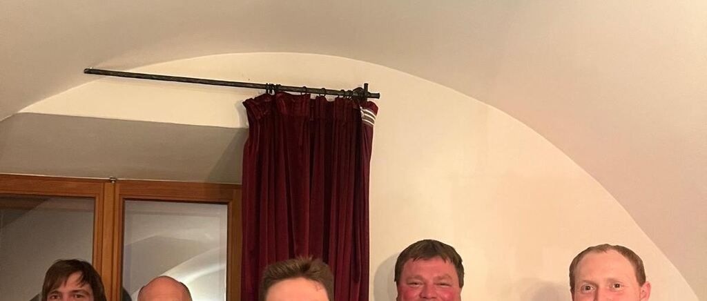 Four men stand together under a curtain. They appear to be smiling and posing for a photo. Behind them is a glass window.