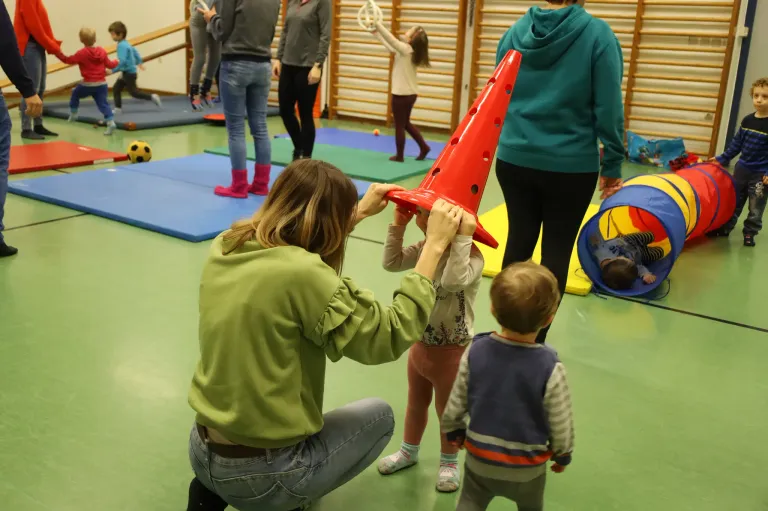 Eine Frau hilft einem Kind, einen roten Kegel auf dem Kopf zu tragen, in einem Spielbereich mit Matten und Spielzeug.