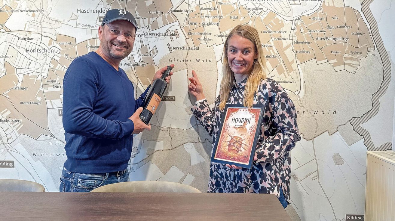 A man and a woman standing in front of a wall map, smiling and posing for a photo. The man holds a bottle of wine and the woman holds a framed award.