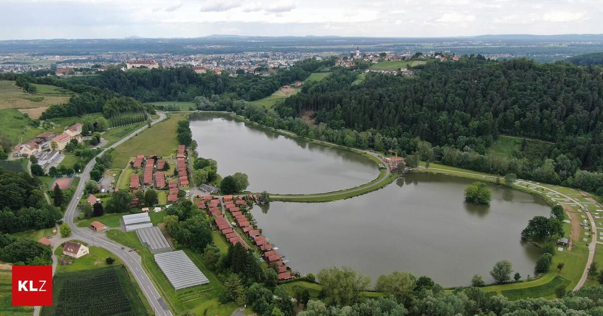 An aerial view of a tranquil lake surrounded by lush greenery, with a cluster of red-roofed houses nearby. In the distance, a town is nestled among trees and mountains.