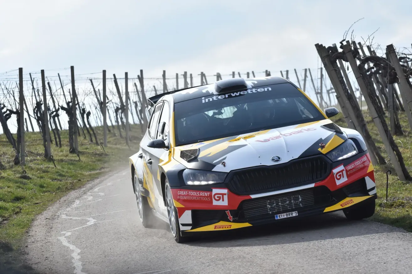 A yellow and black race car is driving on a road with a vineyard and wooden poles in the background. The car has 'interwetten' written on the top and 'GT' on the side.