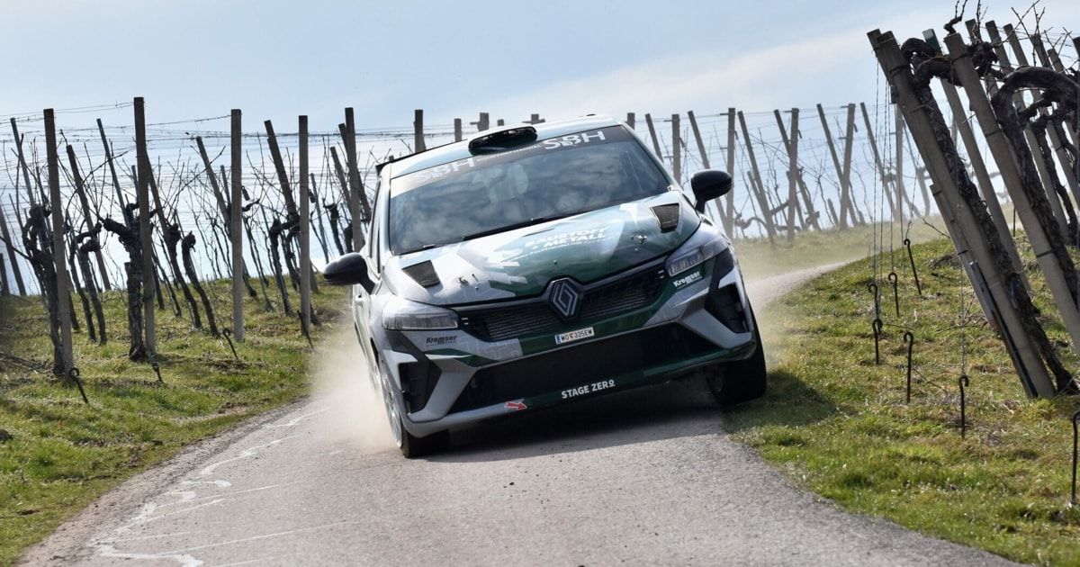 Ein Rallye-Auto fährt auf einer unbefestigten Straße mit einem grün-weißen Farbschema, 'STAGE ZERO' steht auf der vorderen Stoßstange. Dahinter befinden sich Holzzäune und ein Zaun.